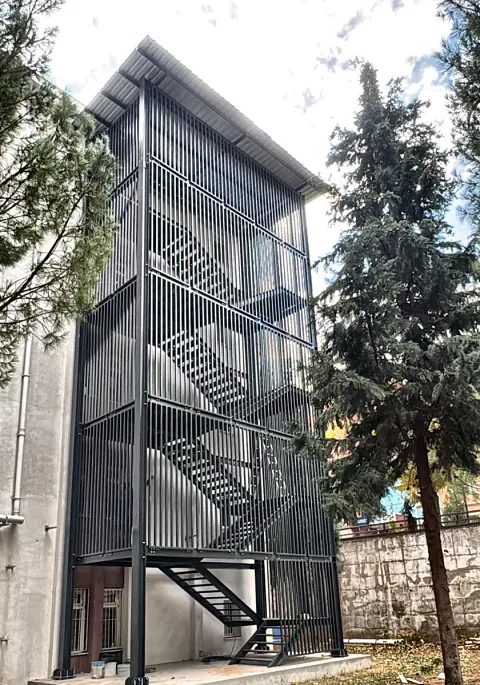 Bahçe tarafında beton zemin üzerine oturan, dikey çubuk kaplamalı çelik merdiven kulesi